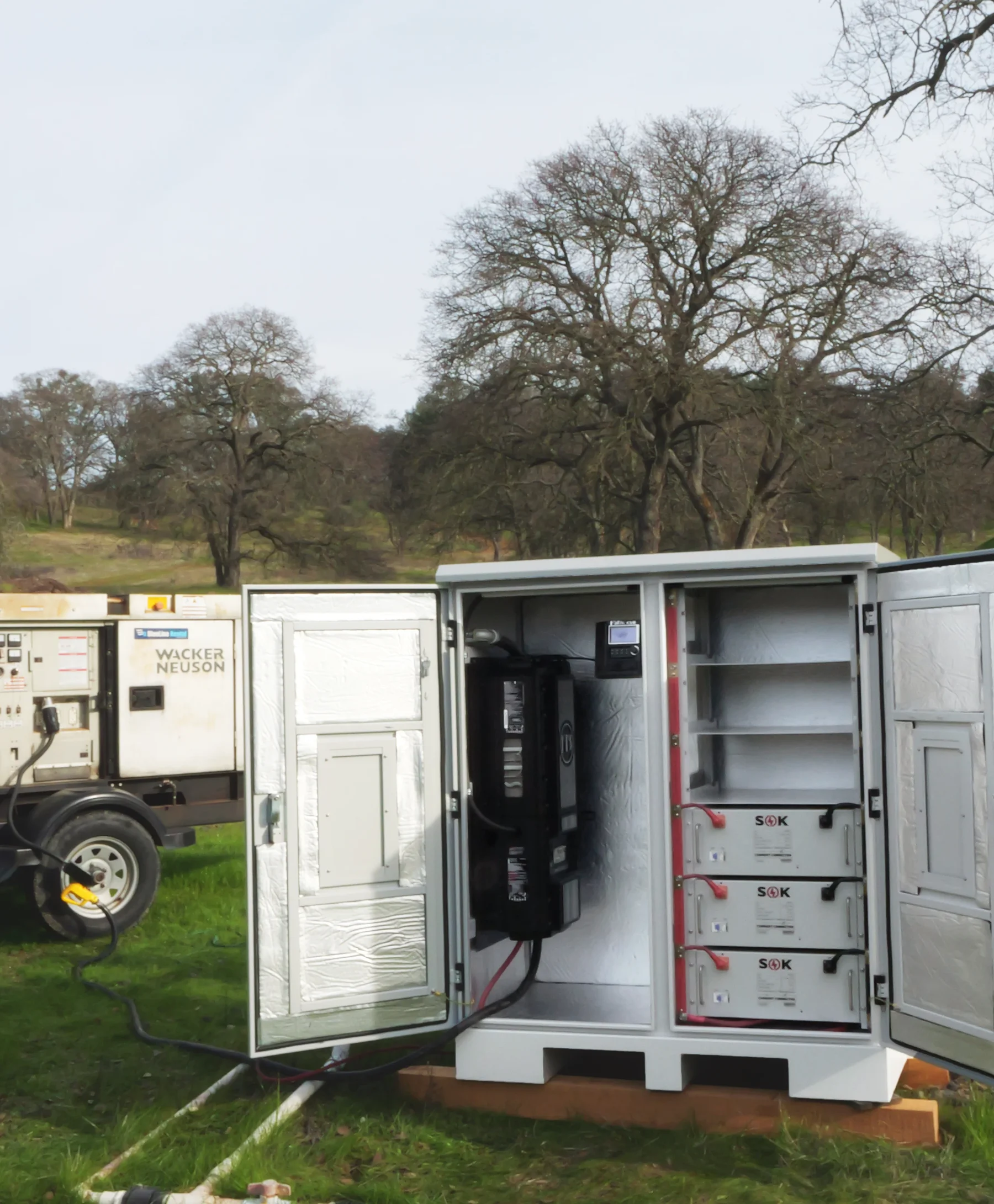 Photo of an inverter and batteries in a cabinet. The cabinet is on a pallet in the middle of a field with trees all around.